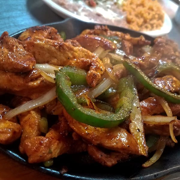Sizzling chicken fajitas with green peppers and onions on a hot skillet, with rice and beans in the background.