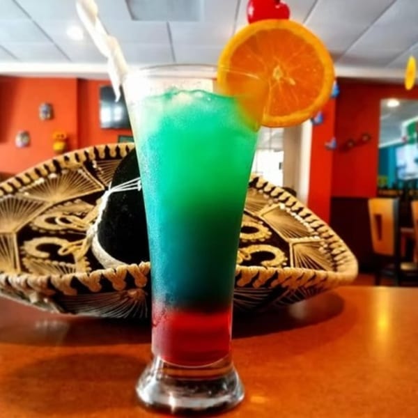 A colorful Mexican flag-themed drink with an orange slice and cherry, next to a sombrero.