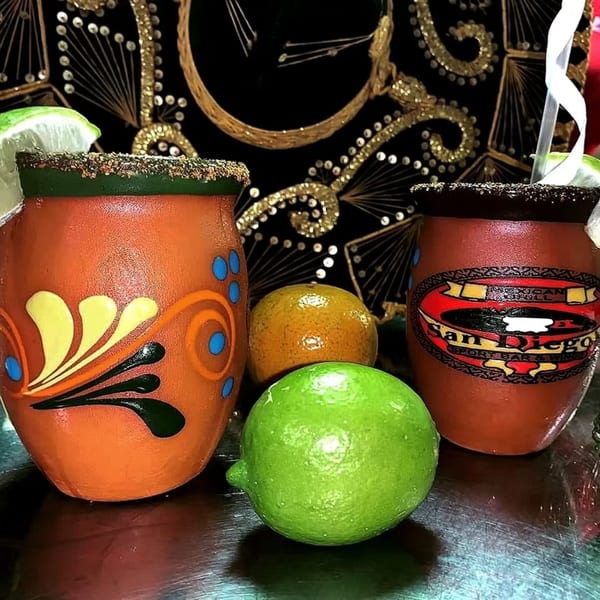 Two San Diego's Mexican Grill margaritas in clay mugs with limes and oranges on a dark surface.