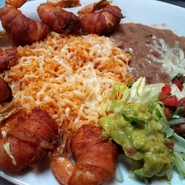 Bacon-wrapped shrimp with Mexican rice, refried beans, guacamole, and salad on a white plate.