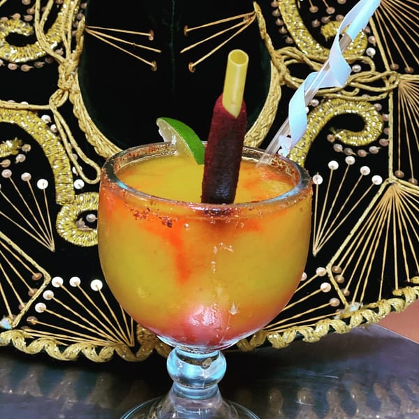 A vibrant yellow Mexican drink with a chili-dusted straw and lime, against a mariachi hat.